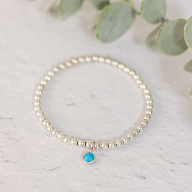 A delicate silver beaded bracelet, the Turquoise December Birthstone Bracelet by Made Here with Love, is centered on a light, textured surface. It features a single turquoise birthstone charm hanging from it. Green leaves are partially visible in the background, adding a natural touch to the composition.