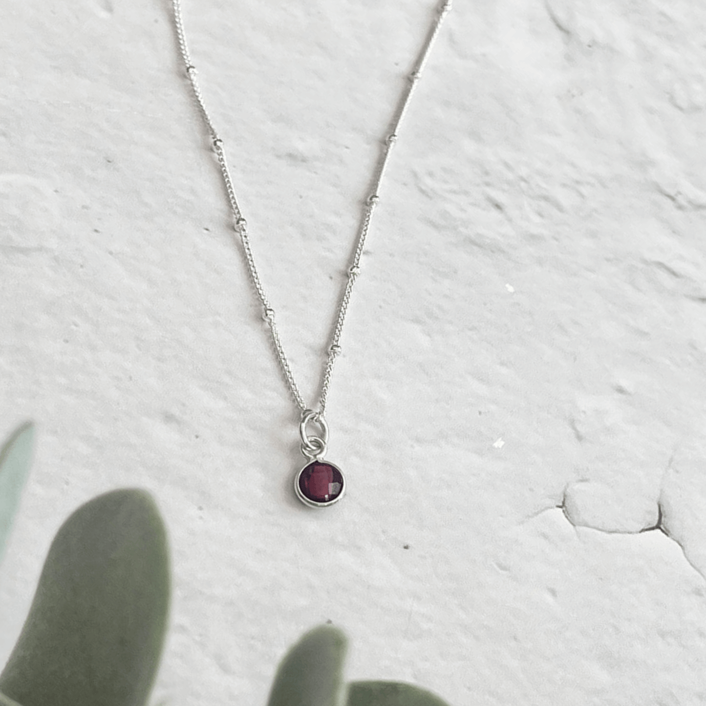 A delicate Silver Garnet Necklace from Made Here with Love, featuring a small, round, deep red gemstone pendant, is pictured against a light, textured background. Green plant leaves blur in the foreground, adding a touch of nature to the minimalist composition.