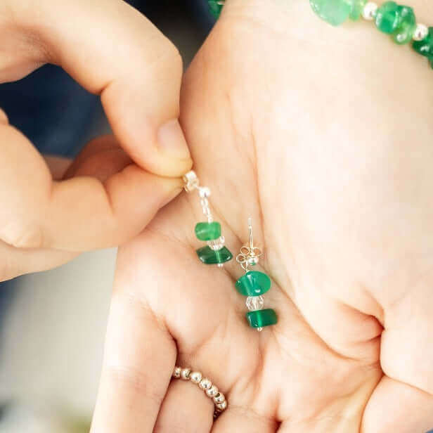 Close-up of a hand holding a pair of Green Agate Drop Earrings by Made Here with Love. The handmade jewelry features three stacked green agate beads with clear bead separators, set on sterling silver hooks. A second hand is adjusting one of the earrings, and the person is wearing a beaded bracelet.