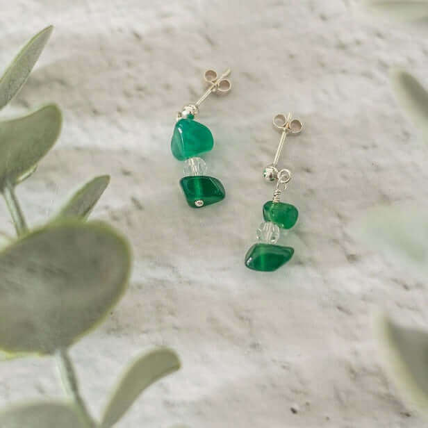 A pair of Green Agate Drop Earrings by Made Here with Love, showcasing small green gemstones and clear beads, set against a soft, white textured background. Green leaves partially frame the image, adding a touch of nature.