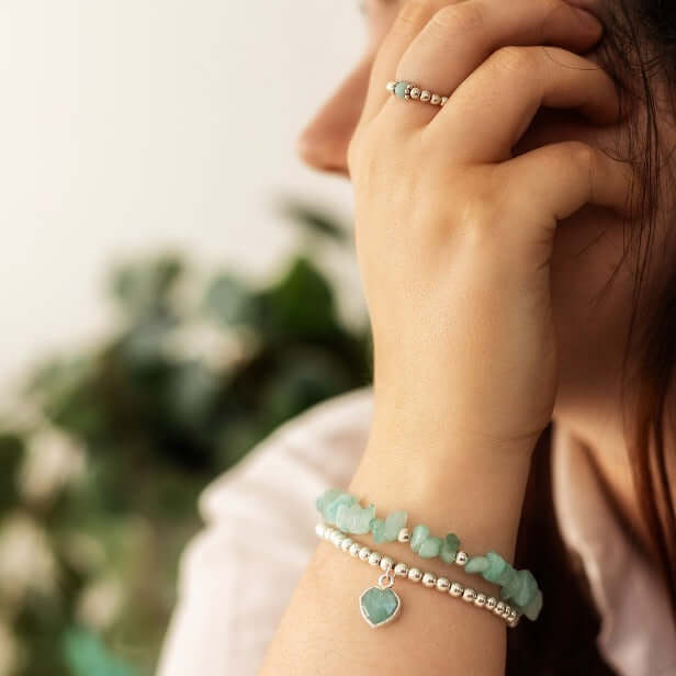 A person with long hair wearing a ring and bracelets adorned with matching green stones, including a Heart Charm Bracelet by Made Here with Love. Sterling silver beads add extra shine as their hand is held up near their face, with a blurred background of greenery.