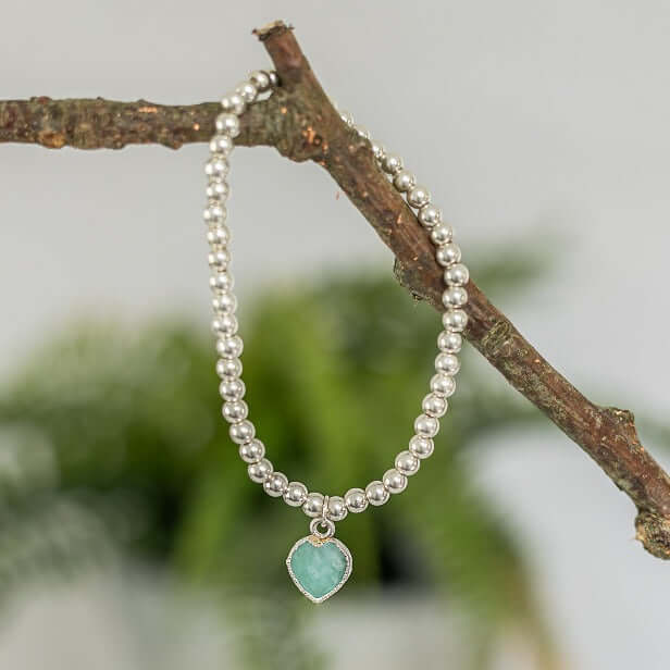 A sterling silver beaded bracelet is draped over a thin tree branch. The bracelet, known as the Heart Charm Bracelet by Made Here with Love, features an Amazonite Heart Charm. The background is blurred, showcasing green foliage.