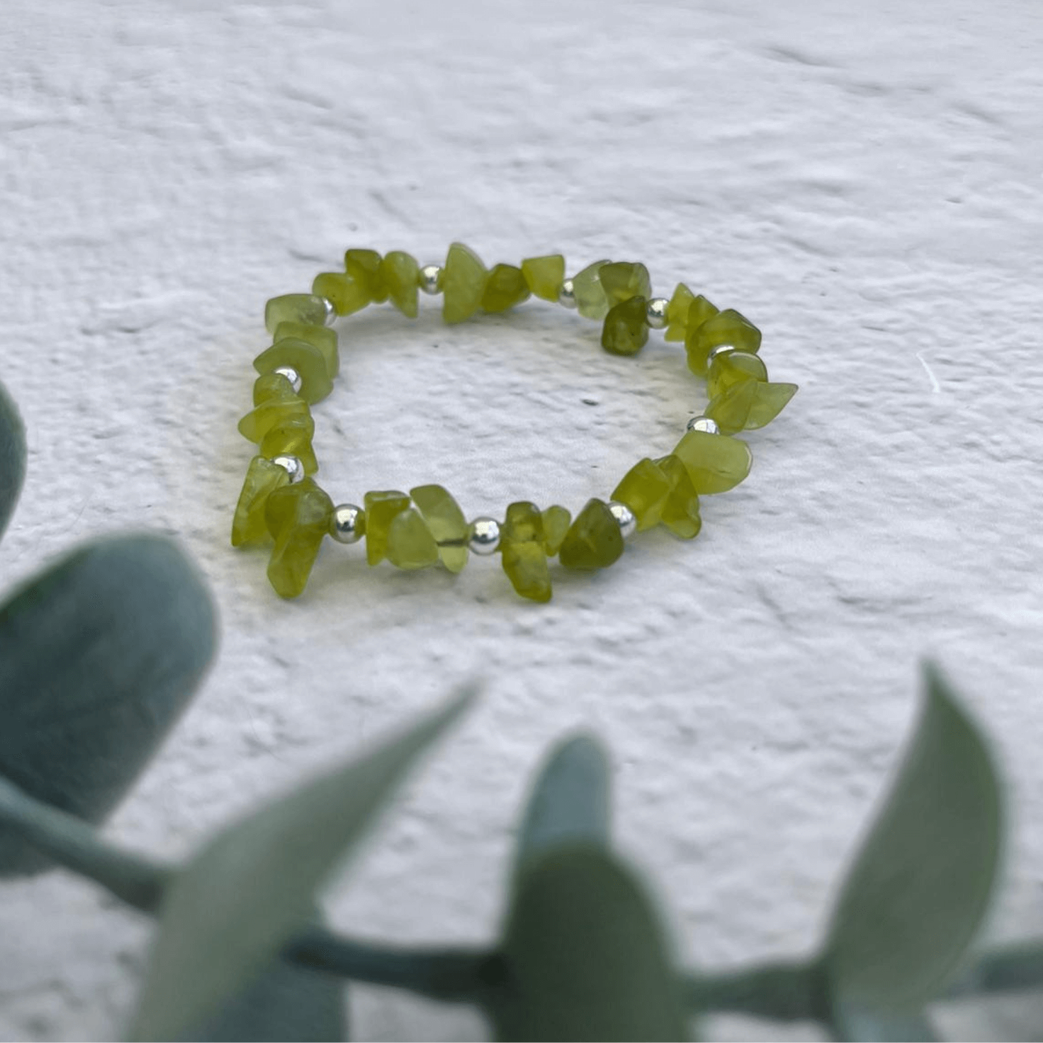 A close-up view of the Made Here with Love Jade Gemstone Bracelet showcases the green jade stones and sterling silver beads, elegantly resting on a textured white surface. Out-of-focus green leaves in the foreground introduce a contrasting element to the composition.