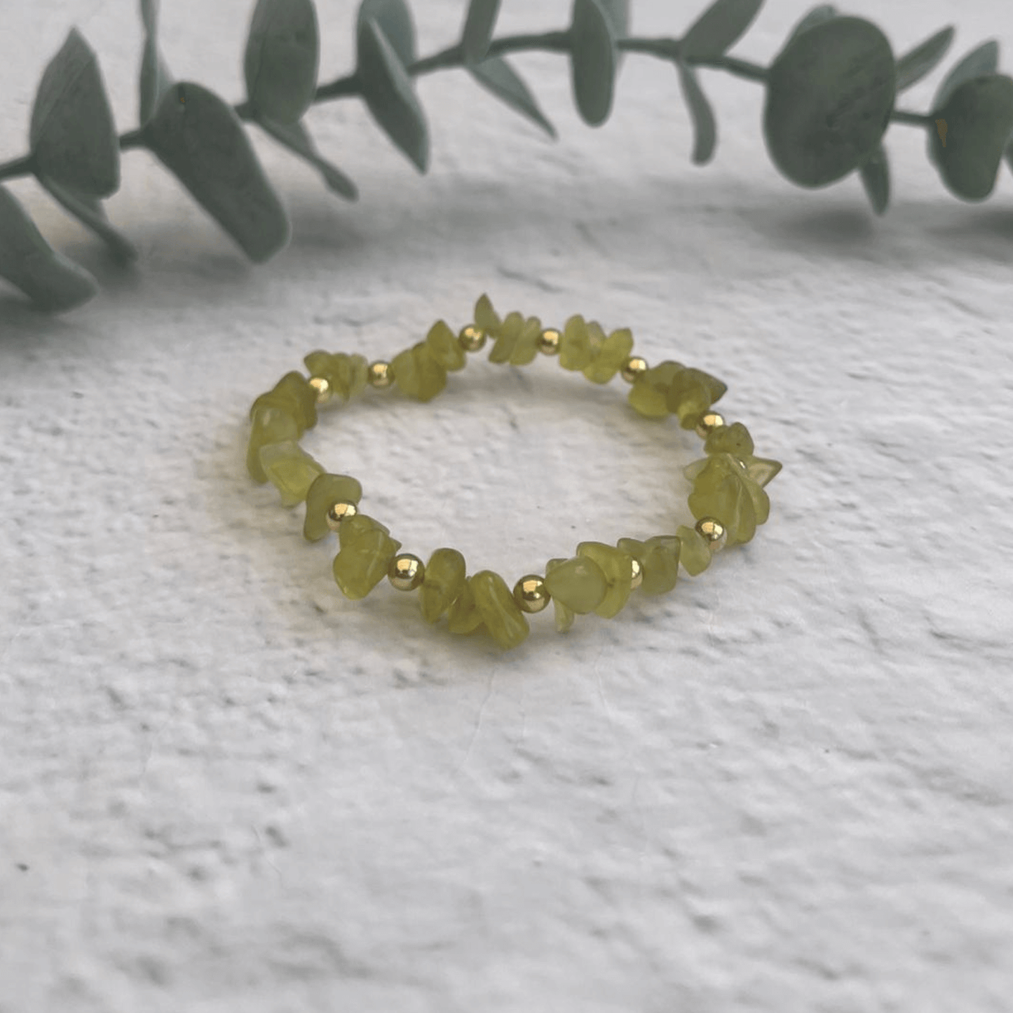 The Jade Gemstone Bracelet by Made Here with Love is crafted from irregularly shaped green stones and small gold-filled beads, set against a textured white surface. A leafy branch adds a natural touch in the background.