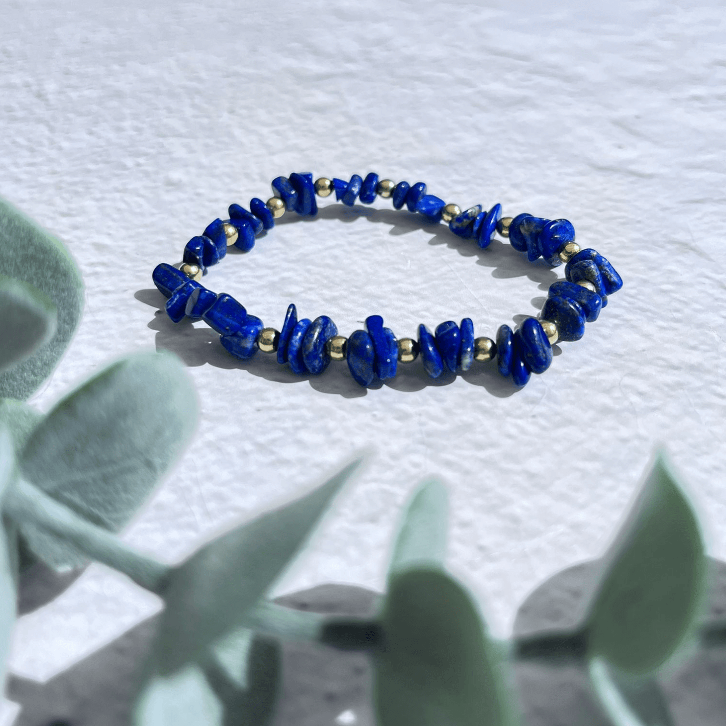 A Lapis Lazuli Stone bracelet from Made Here with Love lies on a textured white surface. The semi-precious gemstone bracelet features irregularly shaped bright blue stones separated by small golden beads. Blurry green leaves are visible in the foreground, providing a contrasting backdrop.