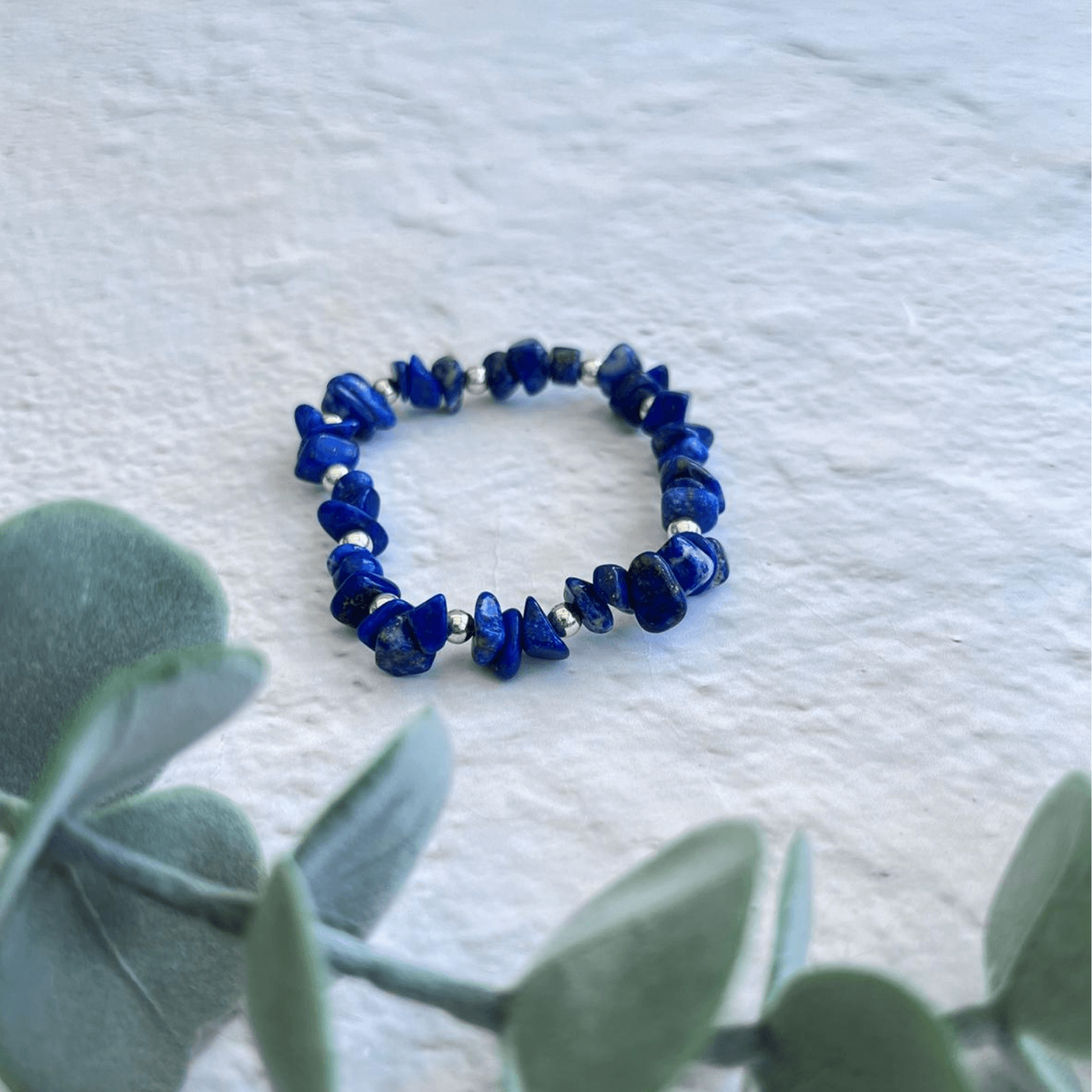 A Made Here with Love Lapis Lazuli Stone bracelet with irregularly shaped stones rests on a light textured surface. Green foliage is partially visible in the foreground on the left, adding a touch of nature to the semi-precious gemstone bracelet.