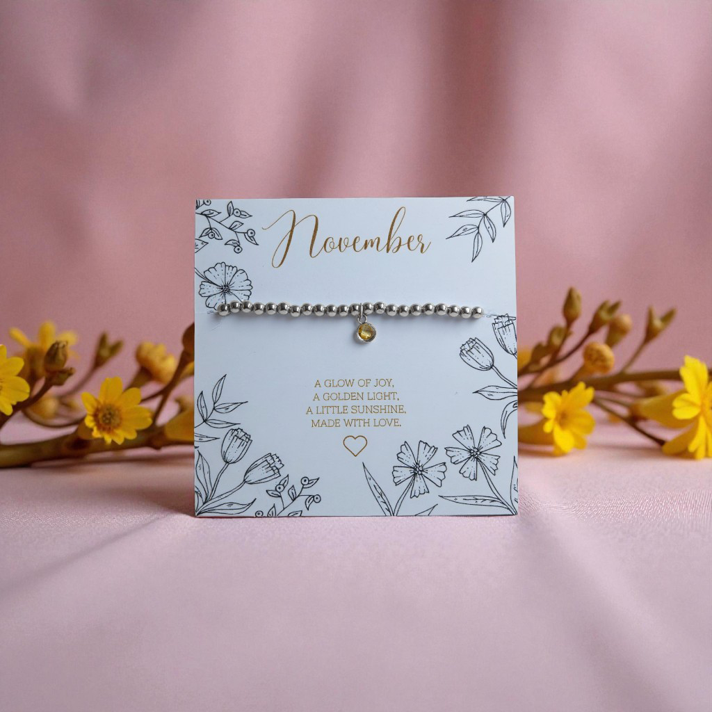 November citrine birthstone bracelet displayed on a floral illustrated card reading ‘A glow of joy, a golden light, a little sunshine. Made with love.’ Surrounded by yellow flowers on a soft pink fabric background.