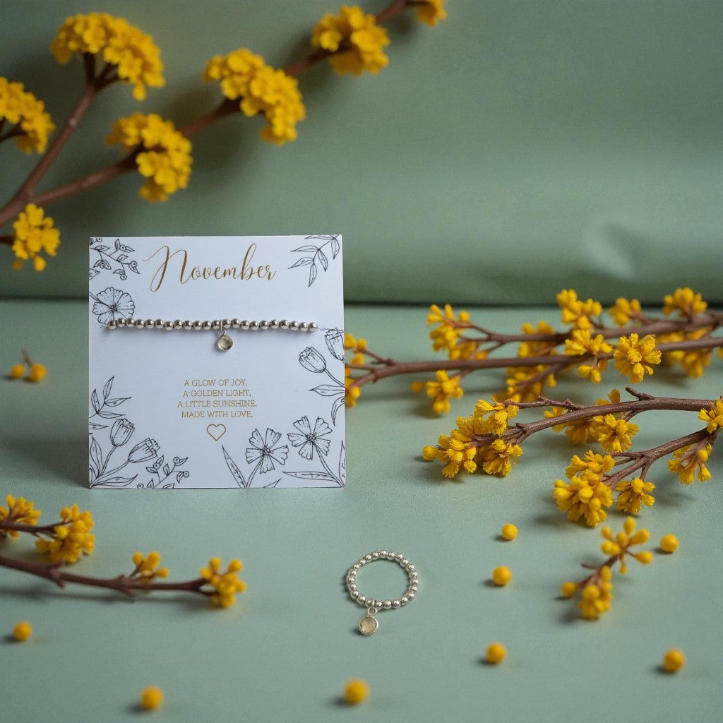 November citrine birthstone bracelet and ring set displayed on a floral illustrated card reading ‘A glow of joy, a golden light, a little sunshine. Made with love.’ Surrounded by yellow flowers and berries on a soft green background