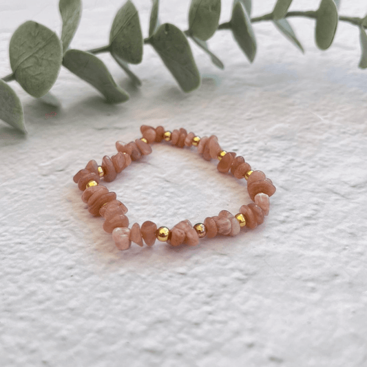 The Peach Moonstone Bracelet from Made Here with Love, featuring a blend of small, irregularly shaped pink stones and tiny gold beads, rests on a light, textured surface. In the background, a sprig of eucalyptus adds a botanical touch to the scene.