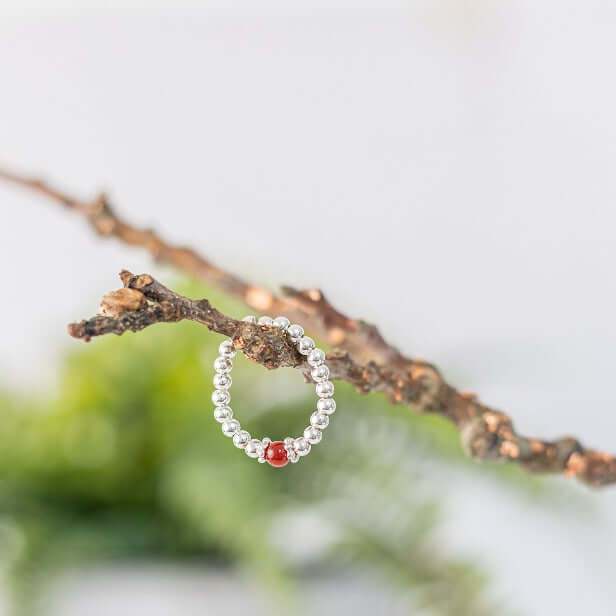 A delicate Red Agate Gemstone Ring by Made Here with Love, crafted in sterling silver with a single red agate gemstone, hangs elegantly from a textured tree branch. The background features soft-focus green foliage, creating a natural and elegant setting for this exquisite jewelry piece.