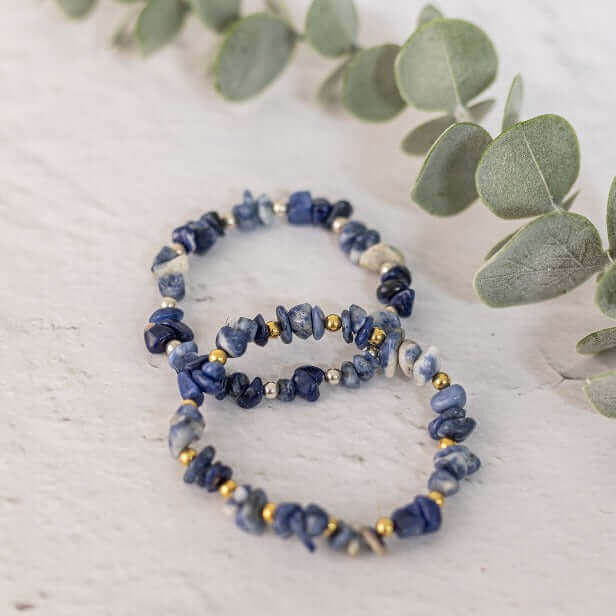 Three Made Here with Love Sodalite Crystal Bracelets, crafted from irregularly shaped blue stones and small gold beads, are displayed on a light surface. Sprigs of green eucalyptus leaves frame the bracelets, adding a natural touch to the composition.