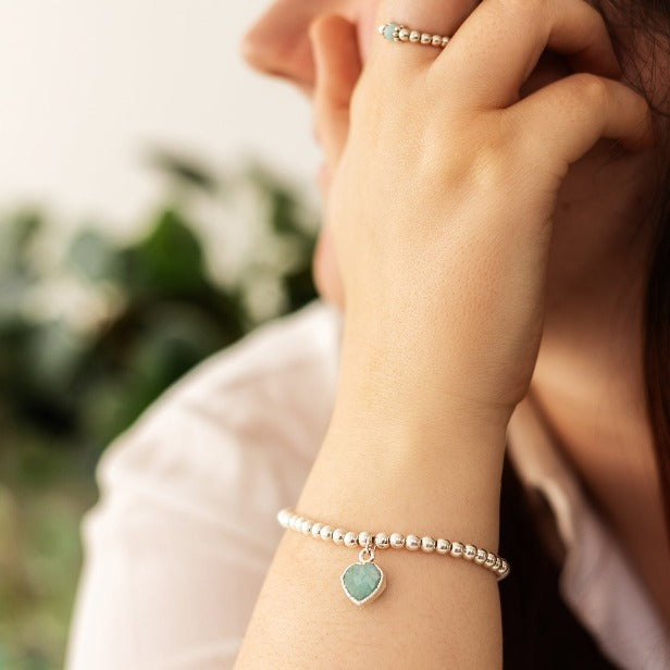 A person with long brown hair rests their hand on their cheek, showcasing a **Made Here with Love Heart Charm Bracelet** with a green amazonite heart charm and sterling silver beads. The background is blurred with greenery. The person wears a white top with short sleeves.