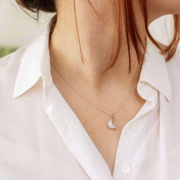 A close-up photo captures a person wearing a white collared shirt and the Made Here with Love Moon Agate Charm Necklace, featuring a delicate crescent moon pendant. The person's red hair is partially visible, and the background is blurred. The necklace, reminiscent of White Moon Agate, adds an elegant touch to their attire.