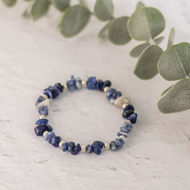 The Sodalite Crystal Bracelet by Made Here with Love, showcasing irregularly shaped blue sodalite stones paired with small silver beads, is elegantly displayed on a white surface. Green eucalyptus leaves in the background add a natural touch to the composition.