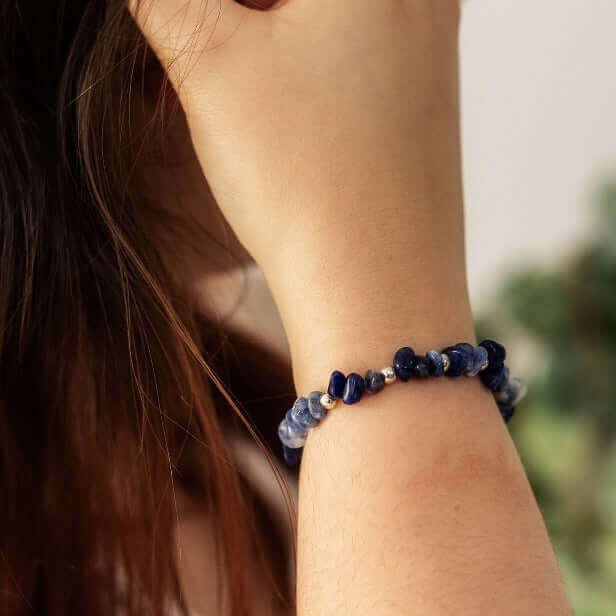 A person with long, dark hair is holding their hand up beside their face, wearing a Sodalite Crystal Bracelet from Made Here with Love on their wrist. The background is blurred, with hints of green, suggesting an outdoor setting.