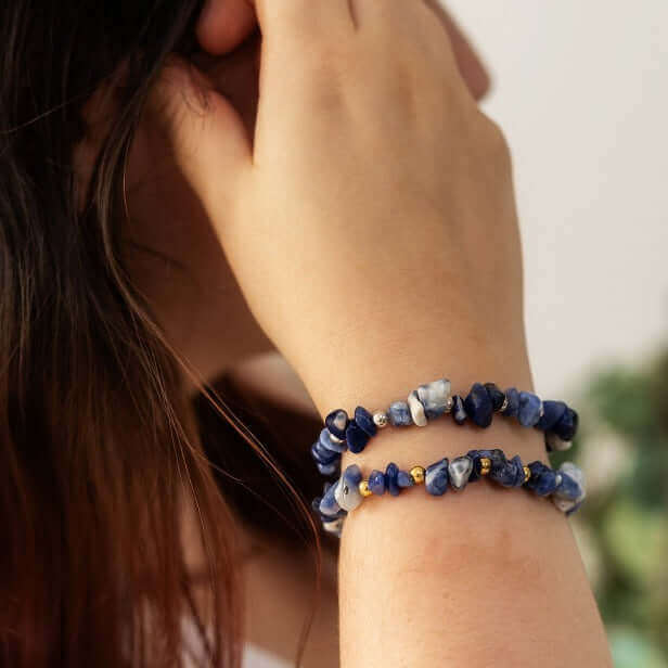 A person with long dark hair is shown wearing two blue beaded bracelets on their wrist, one of which is a Sodalite Crystal Bracelet by Made Here with Love. The person's hand is positioned near their face, and the background is softly blurred, focusing attention on the bracelets.