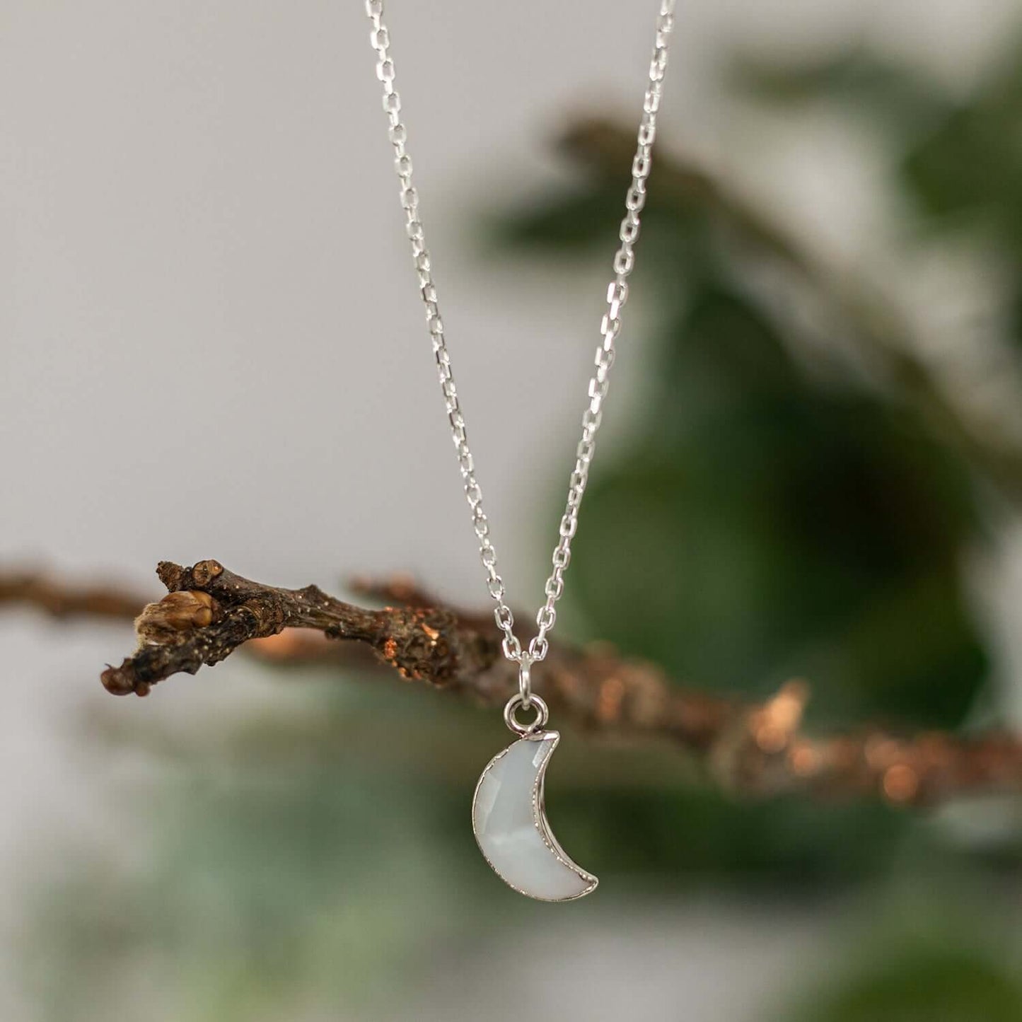 The Moon Agate Charm Necklace by Made Here with Love showcases a delicate crescent moon pendant crafted from White Moon Agate, elegantly suspended from a fine chain. The background softly blurs with touches of greenery and a textured branch, highlighting the simplicity and elegance of this jewelry piece, making it an ideal addition to any gift box.