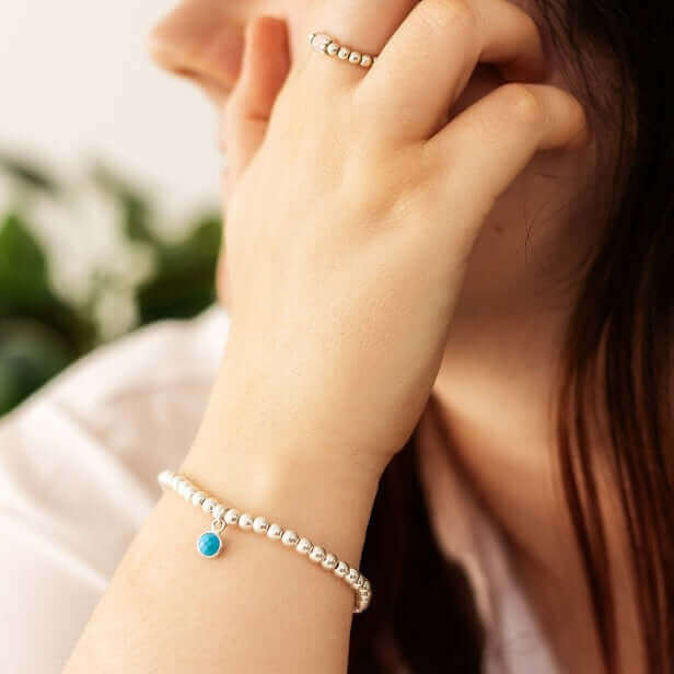 A person with long brown hair is seen from the side, resting their hand on their face. They are wearing a Made Here with Love Turquoise December Birthstone Bracelet featuring a turquoise charm and a matching silver ring. There is a blurred green plant in the background.