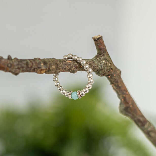 A delicate Amazonite Gemstone Ring by Made Here with Love, featuring a sterling silver beaded band and a single turquoise bead, is perched on a brown branch against an out-of-focus natural green background.