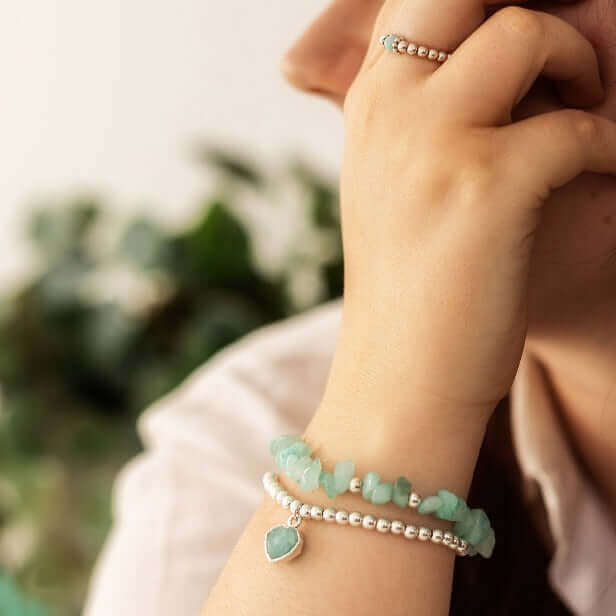 A person rests their chin on their hand, wearing a white bracelet adorned with pearls and green beads, along with matching green bead earrings. Their other hand, partially visible, showcases a Made Here with Love Amazonite Gemstone Ring on the middle finger. The background is blurred with greenery.