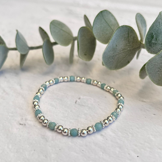 A handmade Amazonite Bracelet by Made Here with Love lies on a flat surface next to a sprig of eucalyptus leaves. The bracelet is composed of alternating small sterling silver and light blue-green beads. The backdrop is a white, slightly textured surface, creating a clean and minimalistic look.