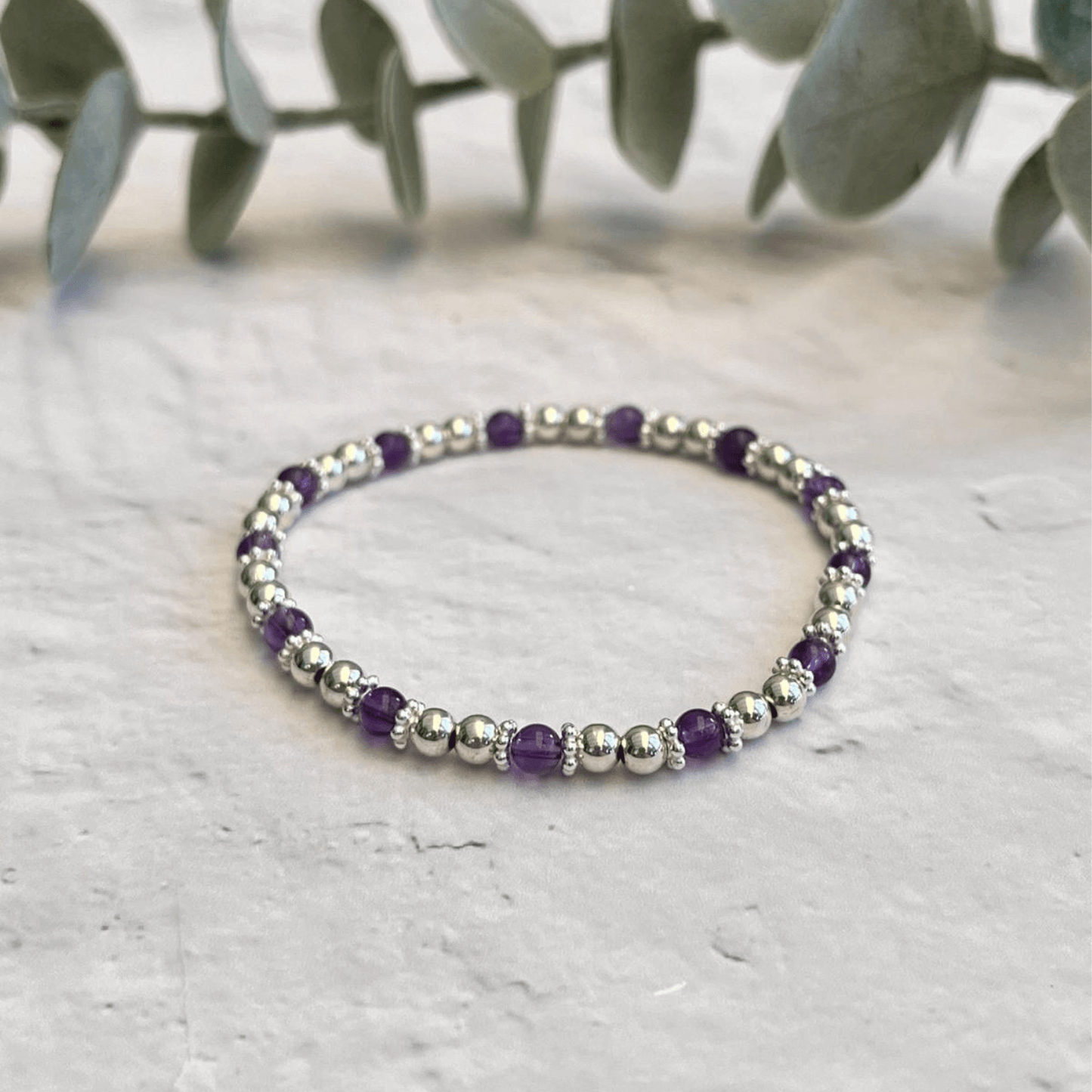 A delicate Dainty Amethyst Bracelet by Made Here with Love sits on a textured white surface, featuring alternating small sterling silver beads and vibrant amethyst gemstones. In the background, blurred green eucalyptus leaves provide a natural backdrop, highlighting this piece of handcrafted jewelry.