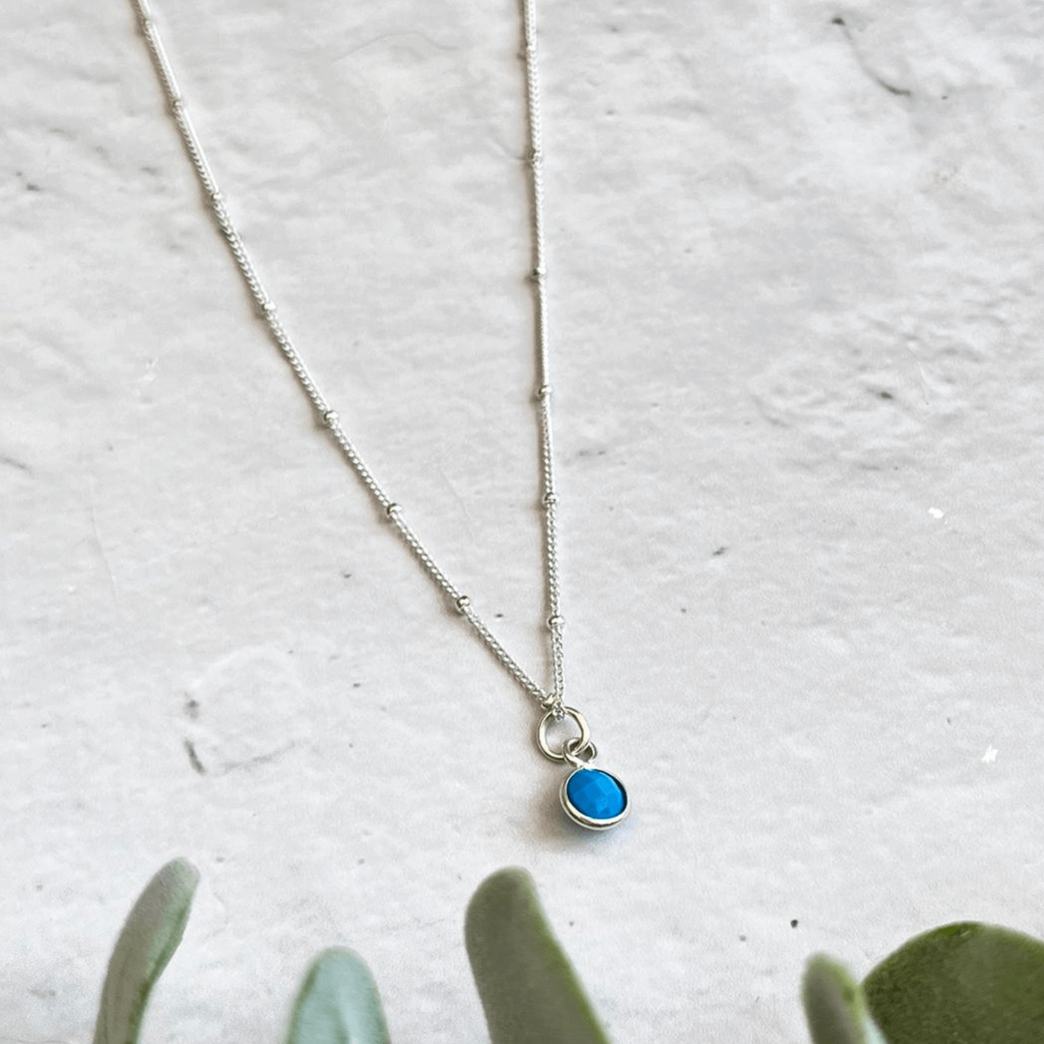A delicate silver necklace with small beads along the chain. The Made Here with Love Turquoise Dec Birthstone Necklace features a blue gemstone pendant encased in silver. The necklace is laid flat on a light-colored, textured surface, with some green foliage partially visible at the bottom.