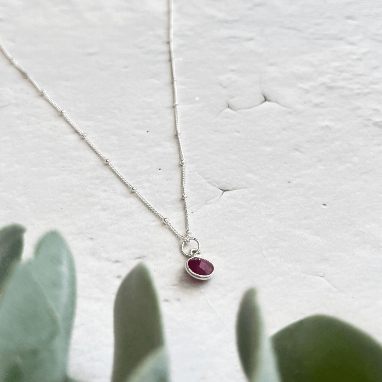 A delicate 925 sterling silver Ruby Birthstone Necklace by Made Here with Love is laid out on a white textured surface. In the foreground, blurred green leaves partially obscure the bottom of the image, adding a touch of natural contrast. This July birthday gift exudes elegance and charm.