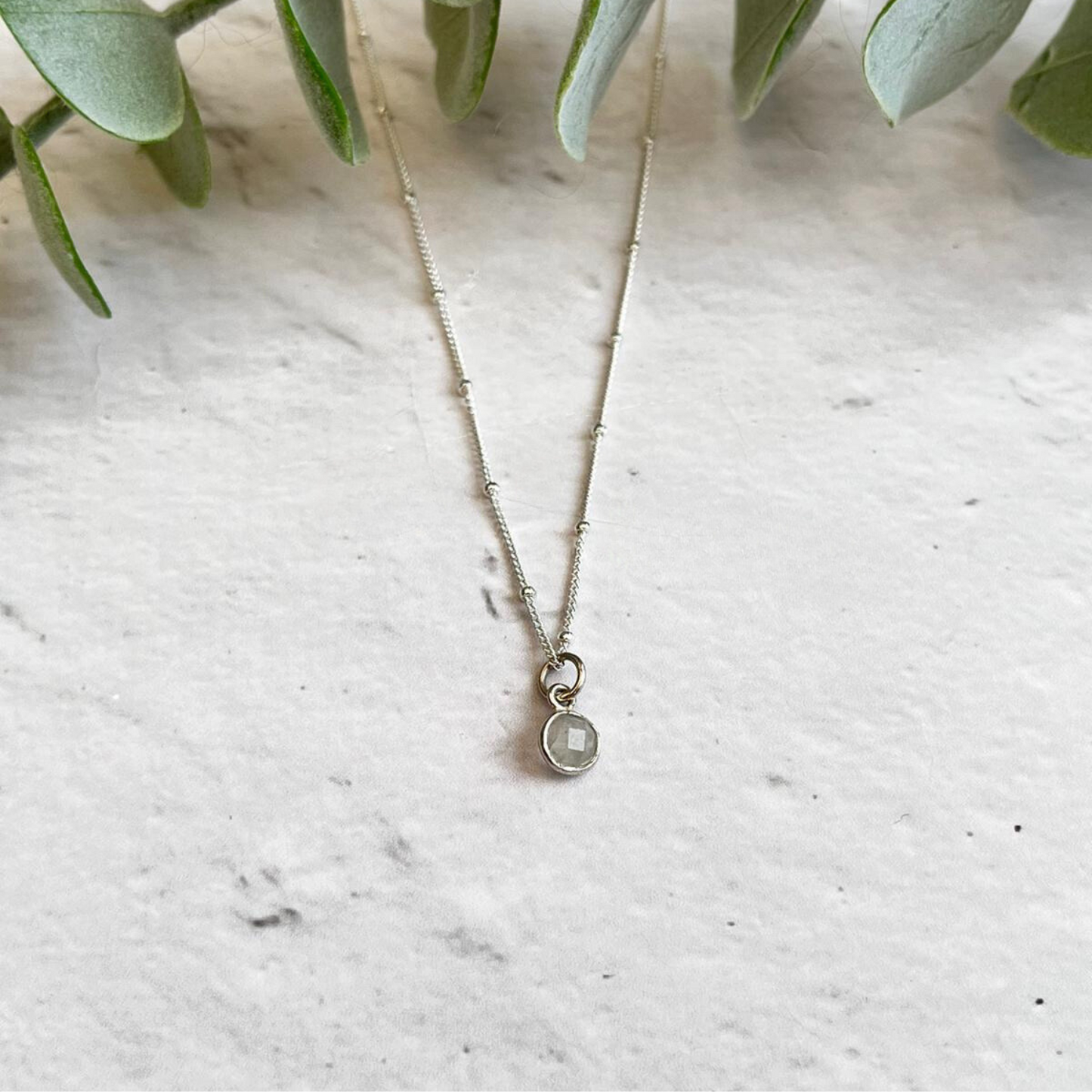 A delicate silver Birthstone Necklace for June by Made Here with Love is placed on a light-colored marble surface. The pendant appears to have a tiny moonstone charm embedded in it, perfect as a June birthday gift. Green leaves frame the top of the image, adding a natural touch.