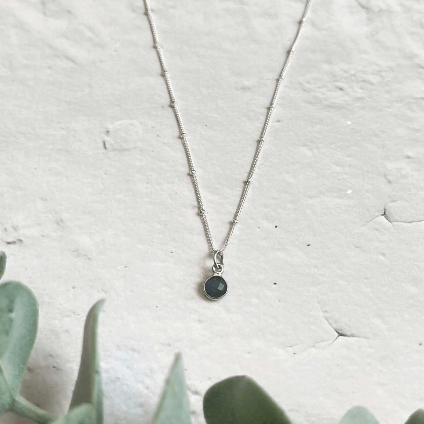 A delicate Birthstone Necklace for March by Made Here with Love with a tiny black circular pendant rests on a light gray, textured surface. A green succulent's leaves can be seen slightly out of focus in the lower left corner of the image.