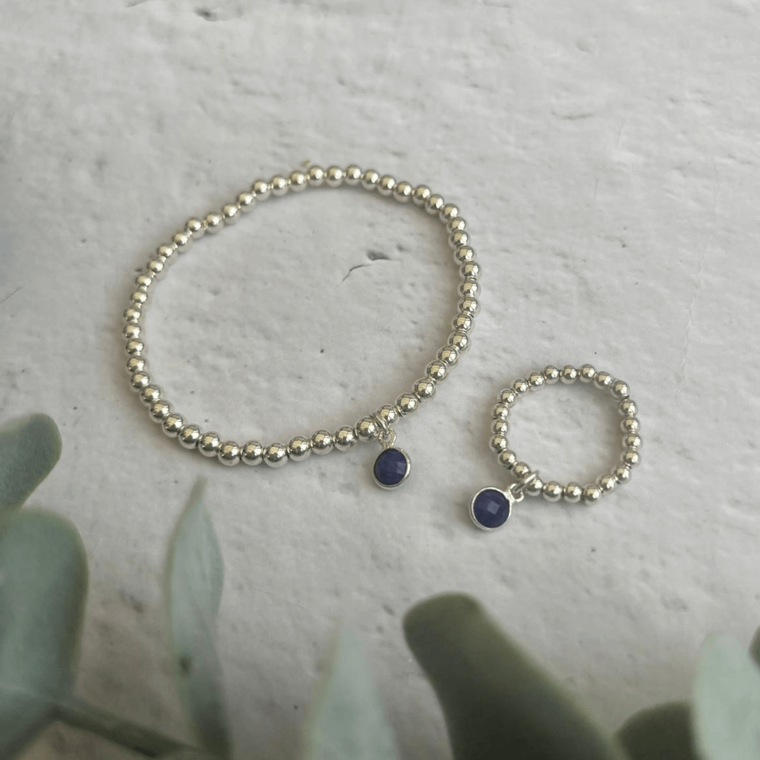 A beaded bracelet and matching ring from the Sapphire Jewellery Sets by Made Here with Love are shown on a light-colored, textured surface. Both pieces are made of small silver beads and have a small circular blue charm. Eucalyptus leaves are partially visible in the bottom left corner of the image.