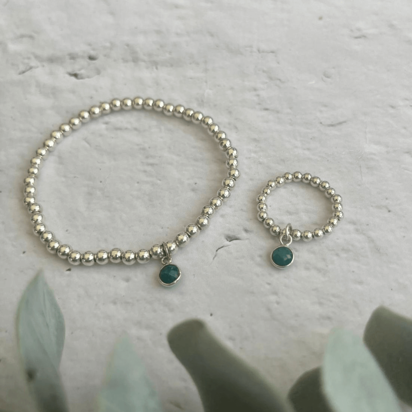 A silver beaded bracelet and matching ring from the Emerald Jewellery Sets by Made Here with Love, each adorned with a small green gemstone charm, are displayed on a gray textured surface. Green leaves are slightly blurred in the foreground, adding a touch of natural elements to the image.