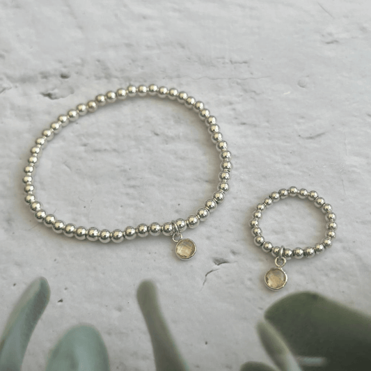 A set of Made Here with Love Citrine November Jewellery Sets on a concrete surface, consisting of a bracelet and a ring. Both are made of small silver beads and feature a single round, light-colored charm. Green succulent leaves are visible in the bottom left corner of the image.