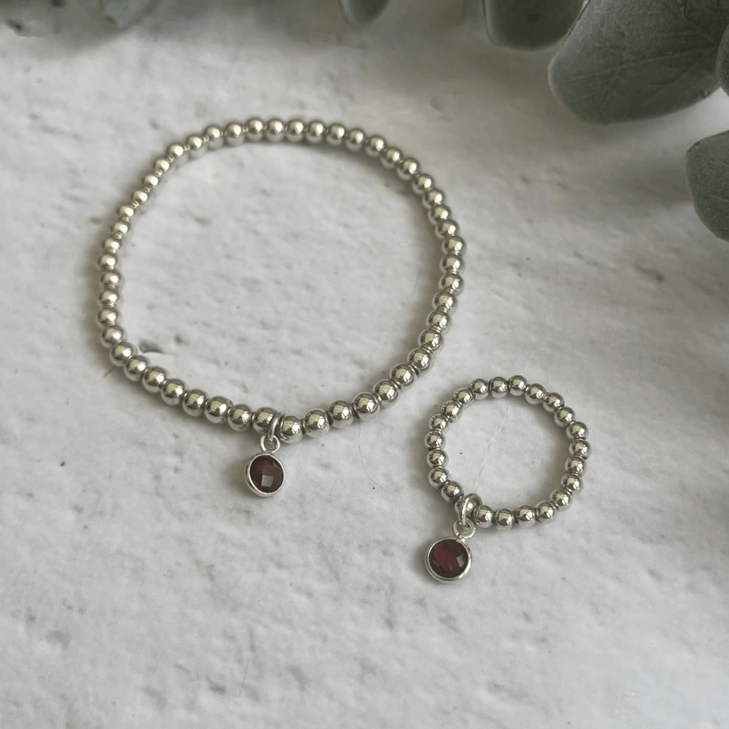 A beaded silver bracelet and ring set with small, round red gemstone charms on both pieces, displayed on a white textured surface with green leafy plants in the background. This is the Garnet Jewellery Sets by Made Here with Love.