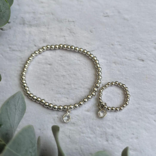 A Clear Quartz April Jewellery Set from Made Here with Love, including a silver beaded bracelet and matching ring featuring Clear Quartz April Birthstone charms, are positioned on a white textured surface. Handcrafted sterling silver beads accentuate their elegance. Greenery partially frames the image on the left side, adding a natural element.