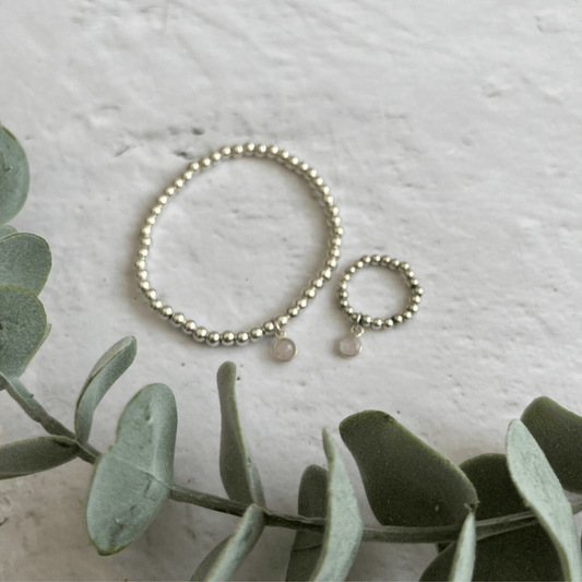 A delicate silver beaded bracelet and matching ring from the Rose Quartz Jewellery Sets by Made Here with Love are placed on a white textured surface. Both pieces feature a small round pink charm. Adjacent to the jewelry is a green eucalyptus branch for decorative effect.