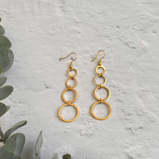 A pair of 24k gold plated, dangling statement earrings featuring a series of interconnected hoops of varying sizes, displayed against a textured white background. Green foliage peeks in from the bottom left corner of the image. These are the Gold Drop Earrings by Made Here with Love.