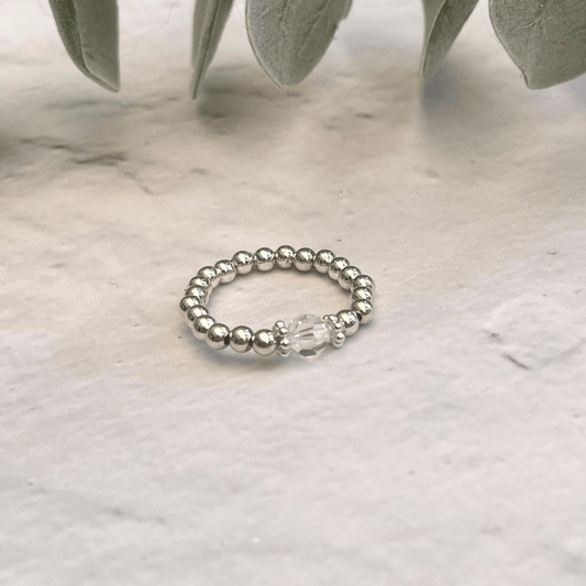 A handcrafted Silver Crystal Ring by Made Here with Love, featuring a single larger Austrian crystal bead at the center, is placed on a light-colored textured surface. Some green leaves are blurred in the background, providing a soft, natural setting.