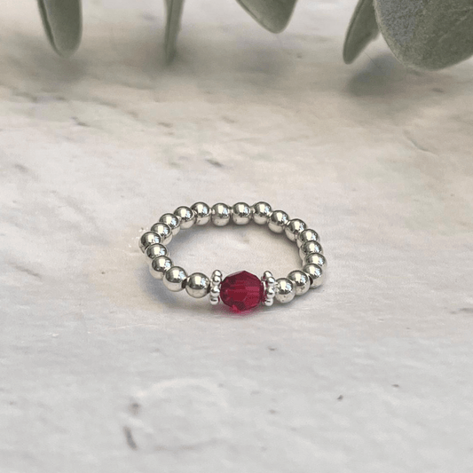 A handcrafted Silver Scarlet Ring by Made Here with Love, adorned with small silver beads and a single red bead at its center, positioned gracefully on a marble surface with green leaves in the background.