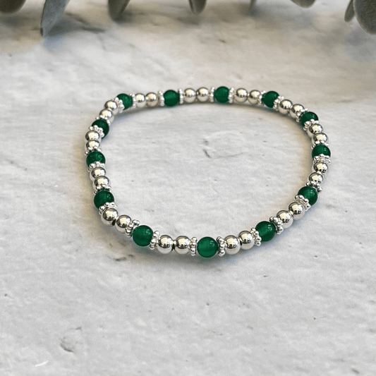 A Made Here with Love Green Agate Gemstone Bracelet with alternating green and sterling silver beads is placed on a light gray, marble-textured surface. The bracelet is circular with a consistent pattern of beads. Some green leaves are faintly visible in the background.