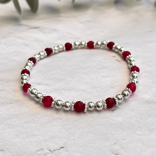 A delicate Pink Agate Gemstone Bracelet by Made Here with Love, made of alternating small sterling silver beads and shimmering red stones, arranged in a simple, elegant pattern. The bracelet is laid out on a light, textured surface with soft-focus greenery in the background.