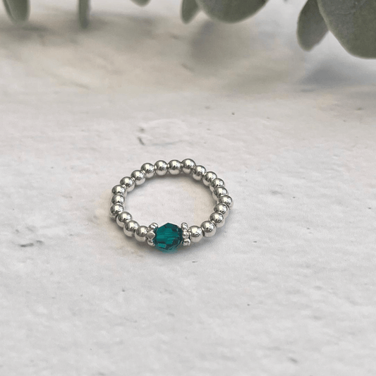 A **Silver Emerald Ring** by **Made Here with Love**, featuring a single green gemstone bead at the center, is placed on a light gray surface. The background includes a few out-of-focus green leaves. This delicate piece of sterling silver jewelry effortlessly captures natural elegance.