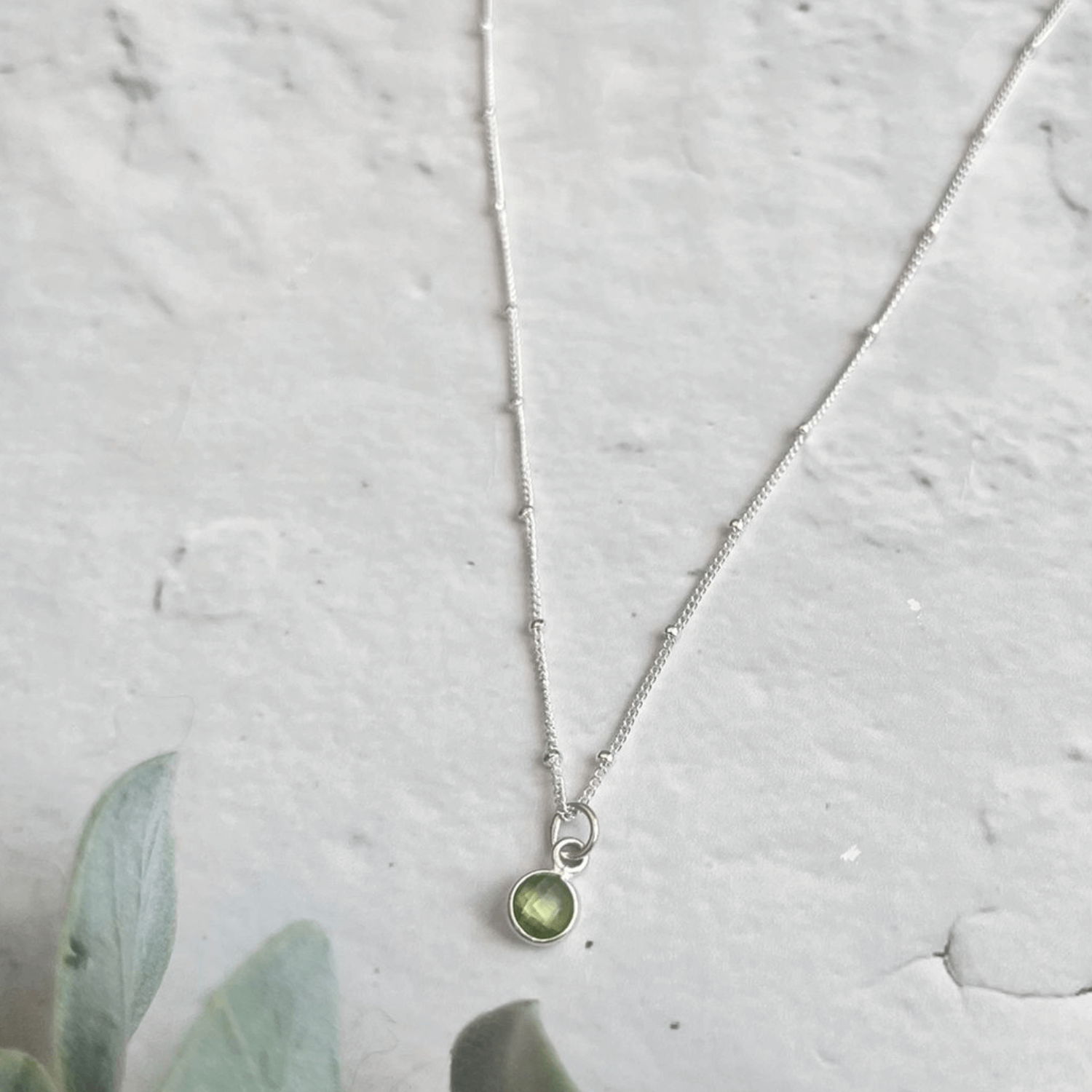 A delicate Made Here with Love Silver Peridot Birthstone Necklace with a small green gemstone pendant rests on a light grey textured surface. Below the necklace, the edge of a green plant can be seen. This beautiful piece is an exquisite example of handmade jewelry, perfect for those celebrating their August birthstone.
