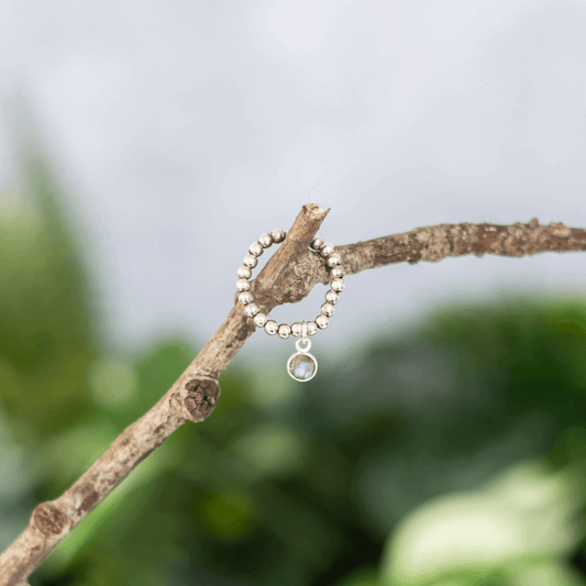 A beaded silver ring with a small clear gemstone pendant hanging from it is placed on a tree branch. The background is blurred, showcasing green leaves and a grayish sky, reminiscent of the elegance found in the Labradorite Birthstone Jewellery Set by Made Here with Love.