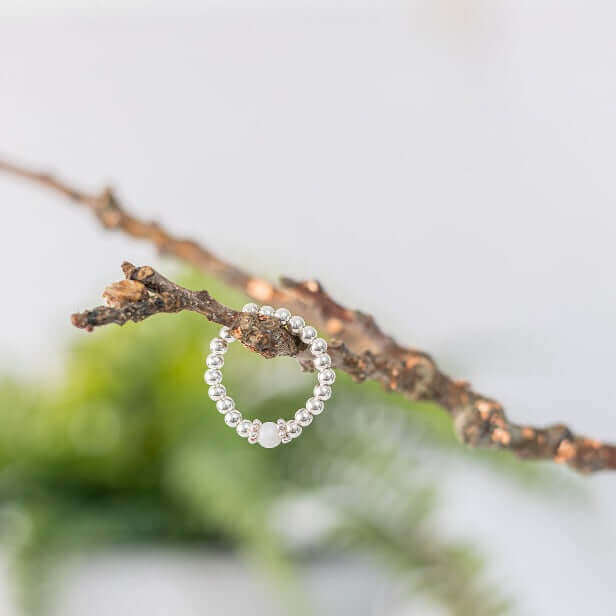 A silver, beaded White Agate Gemstone Ring by Made Here with Love delicately balances on a thin, brown tree branch. The background is softly blurred, featuring hints of green foliage, giving the image a natural and elegant aesthetic.