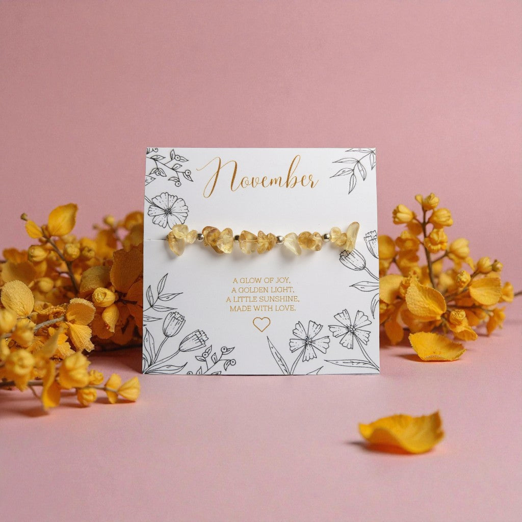 November citrine gemstone bracelet displayed on a floral illustrated card reading ‘A glow of joy, a golden light, a little sunshine. Made with love.’ The card is surrounded by yellow flowers and petals on a soft pink background.