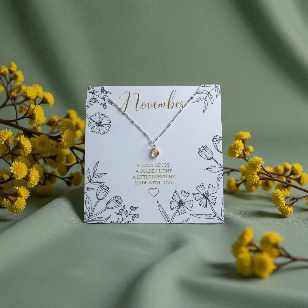 November citrine birthstone necklace displayed on a floral illustrated card reading ‘A glow of joy, a golden light, a little sunshine. Made with love.’ Surrounded by yellow flowers on a soft green fabric background