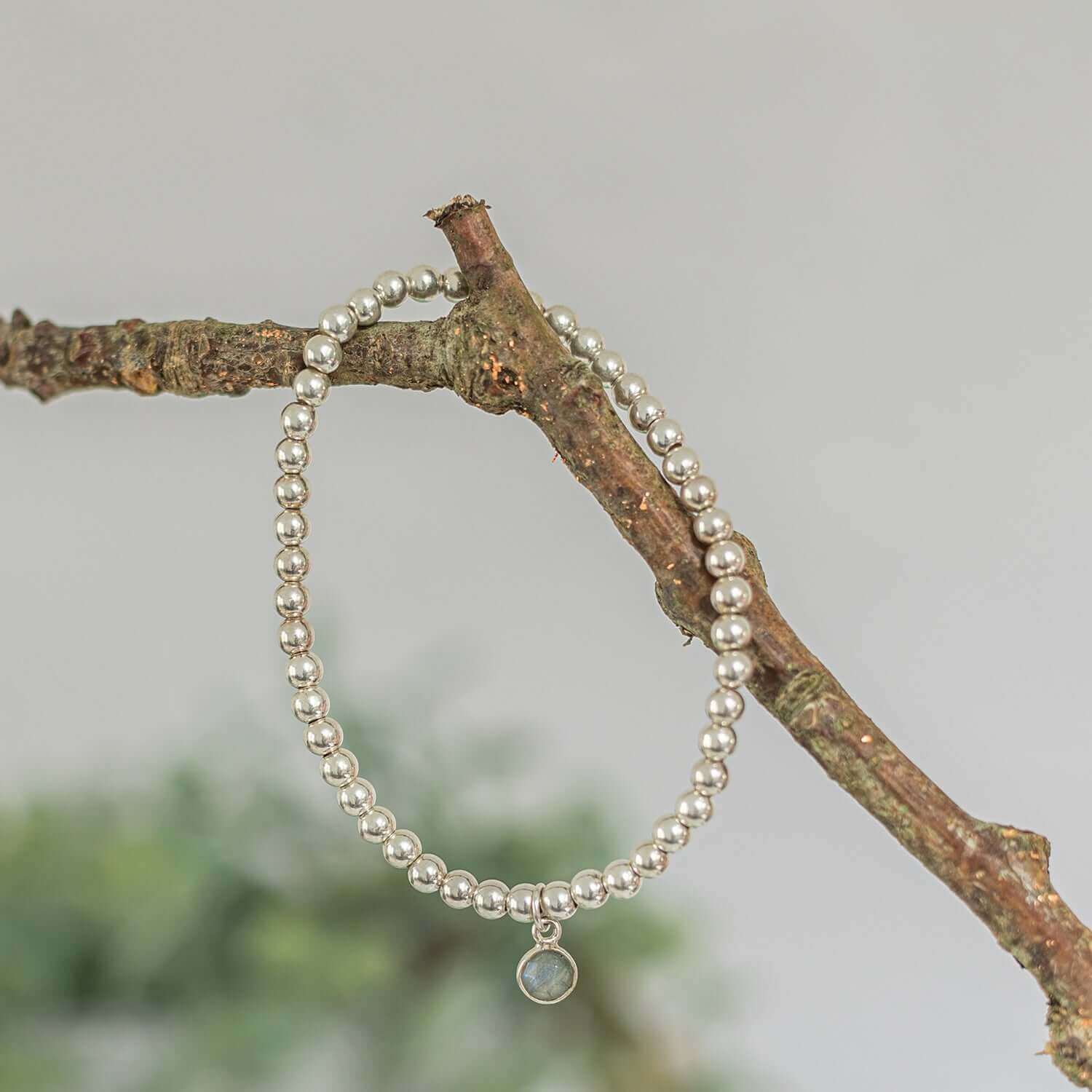 A delicate silver labradorite beaded bracelet with a small, round pendant is draped over a rustic tree branch. The background is blurred, emphasizing the elegant simplicity and natural setting of this Labradorite Birthstone Jewellery Set by Made Here with Love.