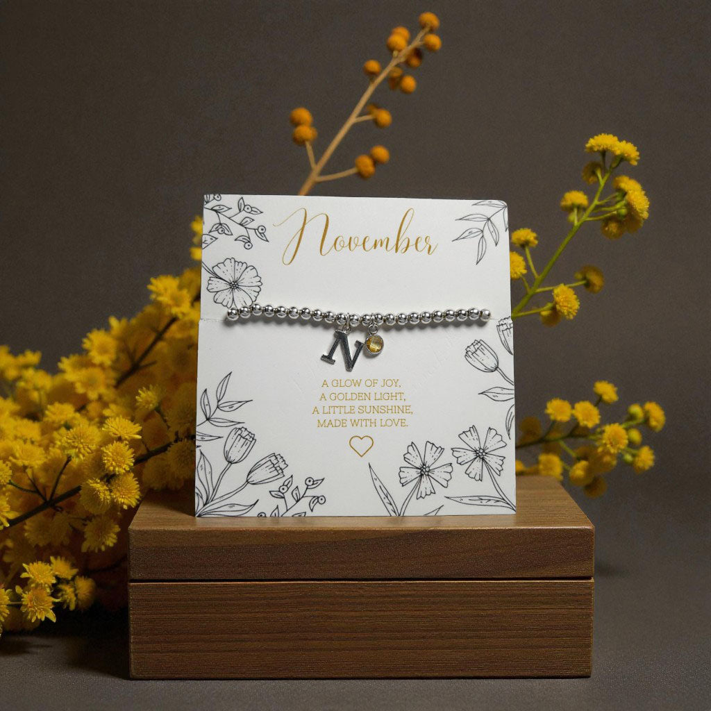 Personalised November citrine birthstone and initial bracelet displayed on a floral illustrated card reading ‘A glow of joy, a golden light, a little sunshine. Made with love.’ Set on a wooden block with yellow flowers and berries against a warm brown background.