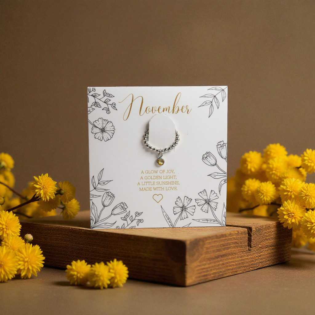 November citrine birthstone ring displayed on a floral illustrated card reading ‘A glow of joy, a golden light, a little sunshine. Made with love.’ Set on a wooden block with yellow flowers against a warm brown background